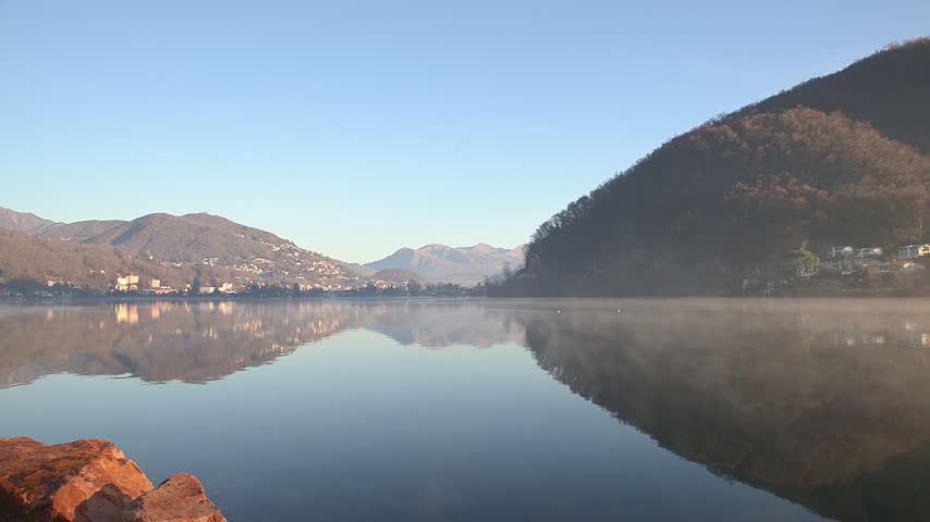 Lake Lugano winter landscape with calm water, fog and mist over the lake, view from Ponte Tresa with Caslano in the background, blue sky and sunny day on the Italy Switzerland border