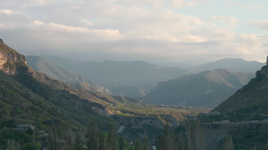 Expansive mountain valley with steep rocky slopes, layered distant ridges, scattered greenery, soft clouds, and warm daylight viewed from high overlook above broad natural terrain