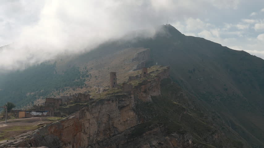 Drifting clouds sweep over rugged mountain ridge with weathered stone ruins, steep cliffs, sparse vegetation, soft daylight, and hazy distant summit in remote scenic landscape