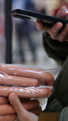 Customer examines packaged sausages while holding smartphone in supermarket aisle. Shopper angles phone over plastic wrapped meat, suggesting product comparison during grocery shopping.
