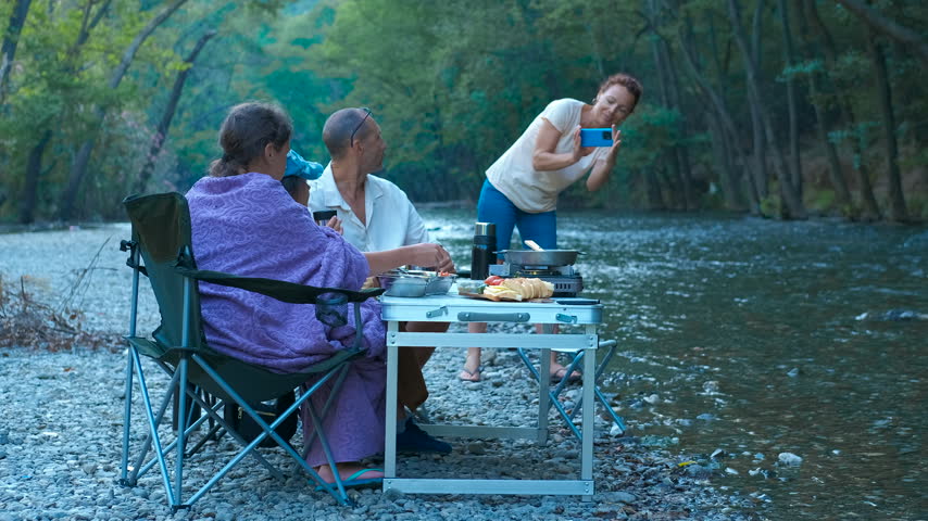 Woman taking pictures of family enjoying picnic by river. Family are enjoying a meal by a river in nature, while one of them takes pictures, capturing memories of their relaxing outdoor picnic