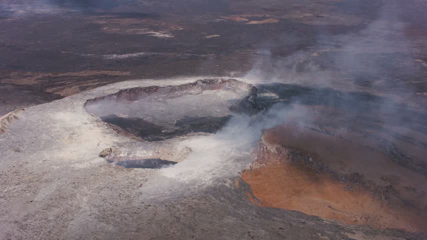 A striking high-angle aerial perspective looking down into a smoking volcanic crater within Hawaii Volcanoes National Park. The footage captures sulfurous white steam and volcanic gases rising from deep vents and hardened lava pits, revealing a vast, desolate surrounding landscape of dark, textured basaltic lava fields and volcanic rock formations under clear daylight.
