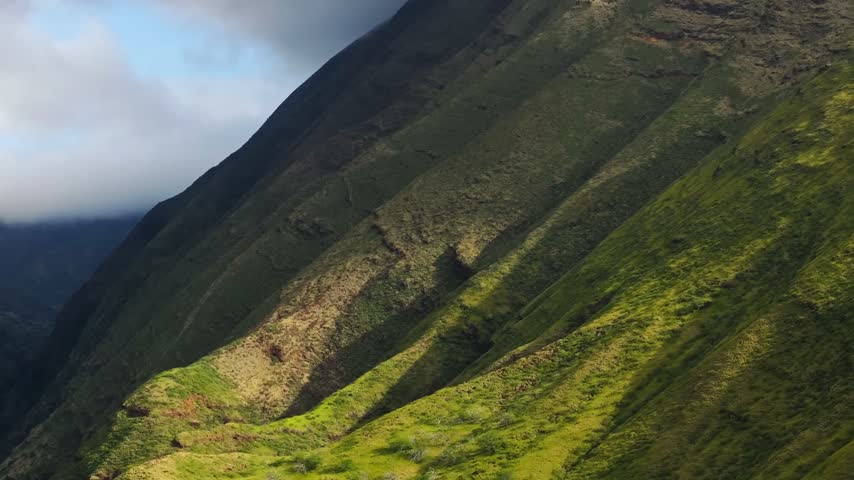 A beautiful high-angle panoramic shot looking across the steep, textured green mountain slopes of Iao Valley in Maui, Hawaii. The footage captures the vibrant mossy ridges and deep valley crevices contrasted by moving shadows from low-hanging white storm clouds passing over the rugged peaks under soft, shifting daylight.
