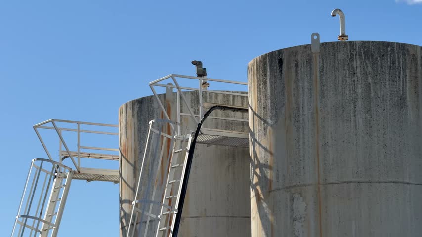 A tilt down with a slow push in on a cylindrical industrial tank with a metal ladder and platform, set against a clear blue sky, highlighting the textures and materials of the abandoned site.