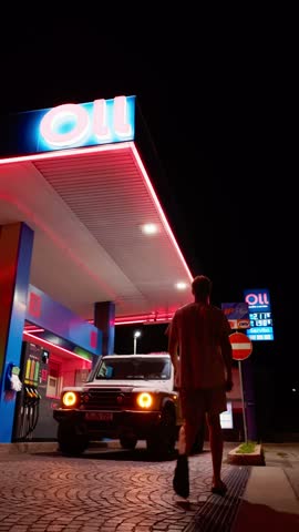 Static vertical hold of man walking to white off roader at neon lit gas station at night, Lago Maggiore, Piedmont, Lombardy, Italian lakes, Italy
