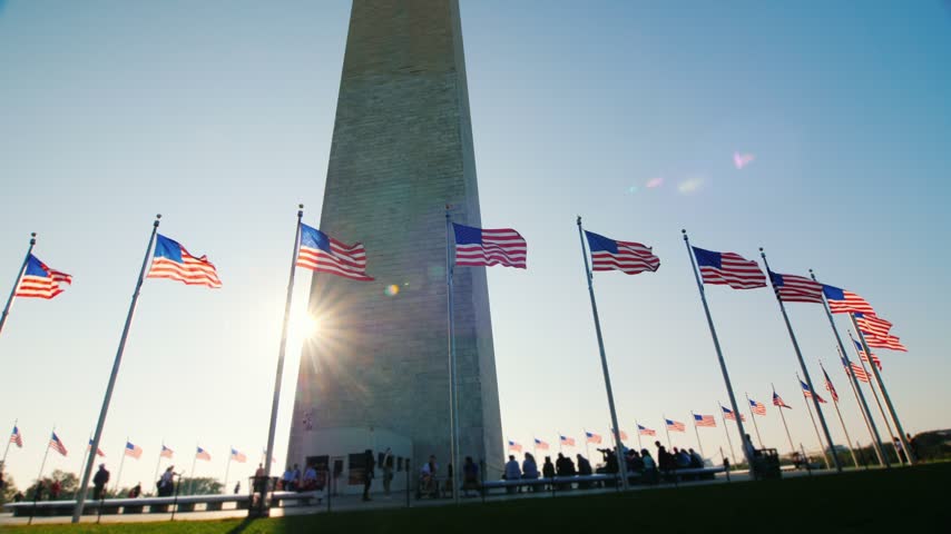 Dynamic footage of marble obelisk landmark with national flags fluttering under clear blue sky during golden hour in Washington DC district.