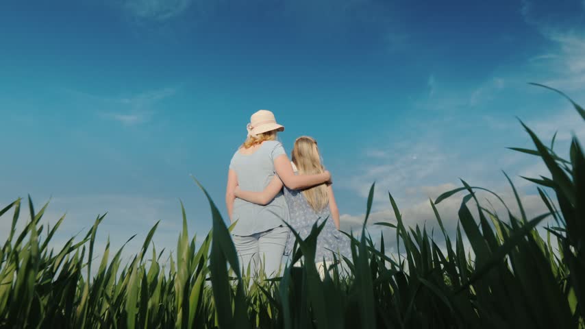 Back view of mother and young daughter hugging while standing in vibrant green agricultural field under bright blue sky during summer day cinematic slow motion footage.