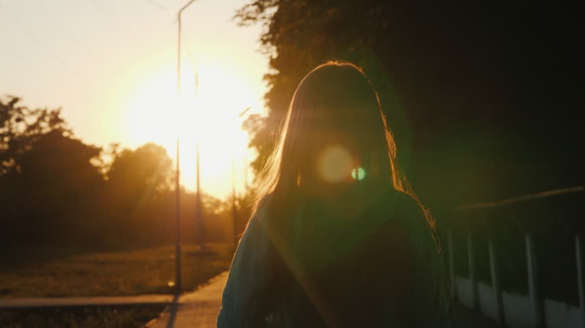Slow motion footage of young girl with long hair spinning and dancing joyfully during sunset in public park. Cinematic backlit shot capturing carefree childhood happiness outdoors.