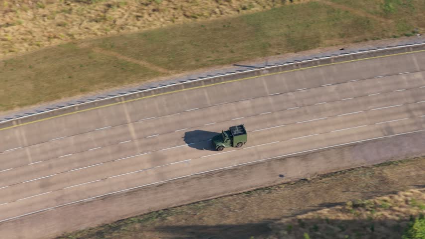 Green military vehicle travels along a wide concrete road at a specialized testing facility while casting long shadows under the bright sun in a dry grassy area