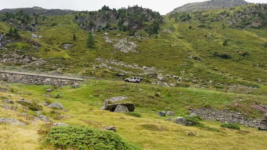 Static wide shot of alpine slope with stone walled pass road and parked SUV above boulder meadow on cloudy day, Dolomites, South Tyrol, Alps, Italy