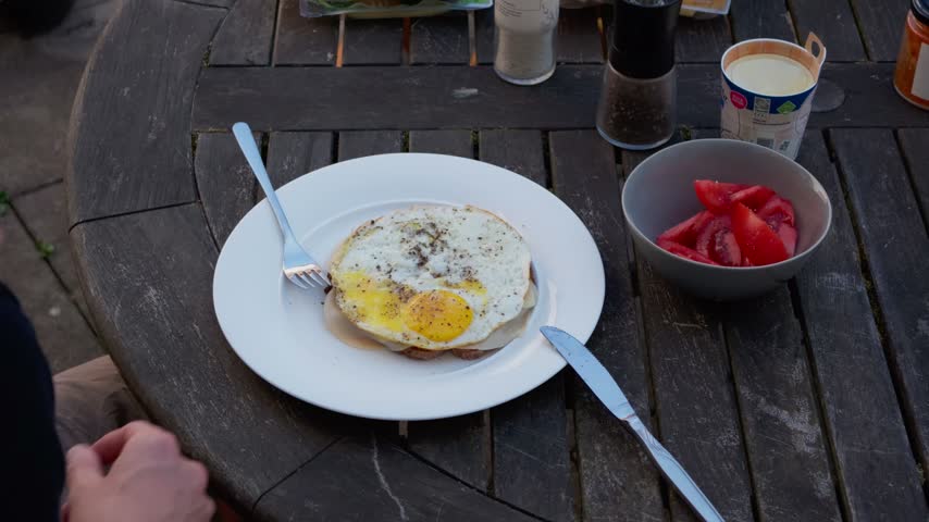 Static close-up of hands cutting a fried egg on toast beside sliced tomatoes on a wooden table in morning light, Scandinavia, Denmark