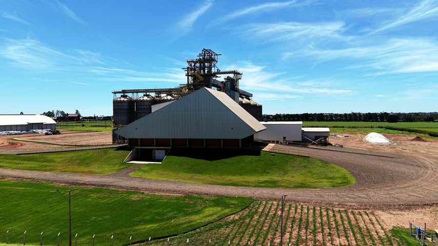 Aerial drone view of a large grain storage and agricultural processing facility with multiple silos, industrial structures and surrounding green farmland under a blue sky.