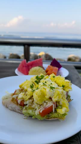 Happy and relaxing morning enjoying scrambled eggs on avocado toast with fresh tropical fruits on a seaside terrace