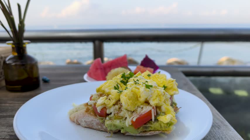 Happy and relaxing morning enjoying scrambled eggs on avocado toast with fresh tropical fruits on a seaside terrace by ocean