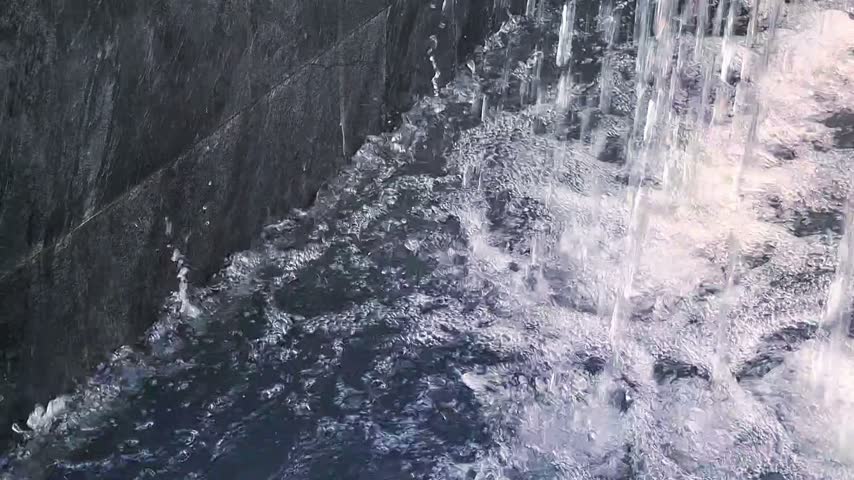 Cascading water curtain falling into stone pool wall creating sparkling ripples and foam bubbles
