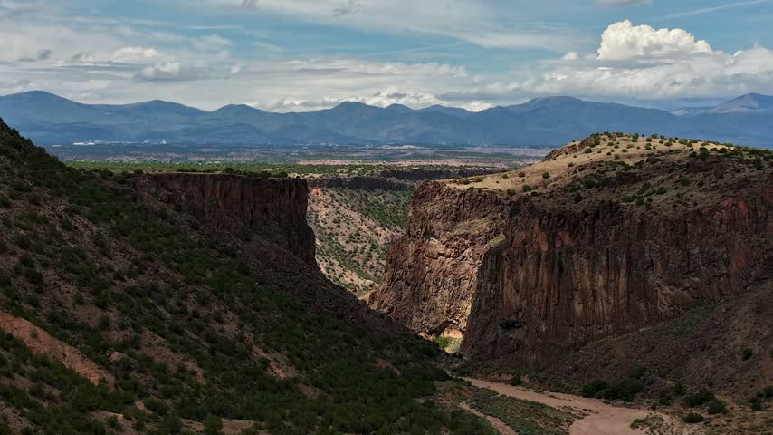 4K drone timelapse flying over Diablo Canyon near Santa Fe, New Mexico. Deep shadows move across the steep basalt cliffs and dry riverbed, with distant blue mountains under a partly cloudy sky.