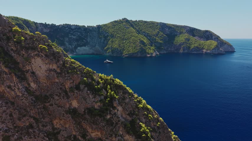 Drone flies forward over a deep blue bay enclosed by pine covered limestone cliffs and a yacht