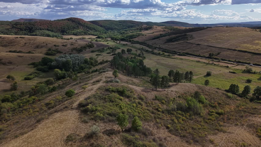 An aerial drone perspective captures the rolling hills and grassy valley of a serene rural landscape under a cloudy sky.