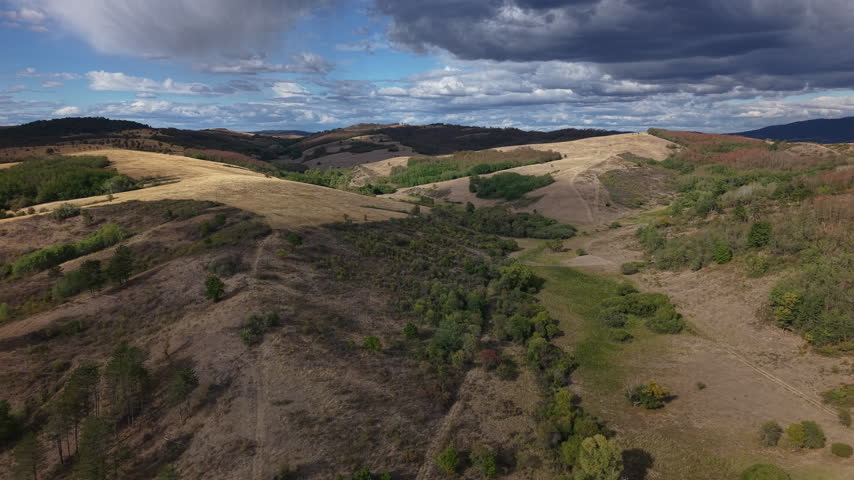 An aerial landscape view of rolling hills with patches of golden autumn grass, green trees, and a dramatic cloudy sky.