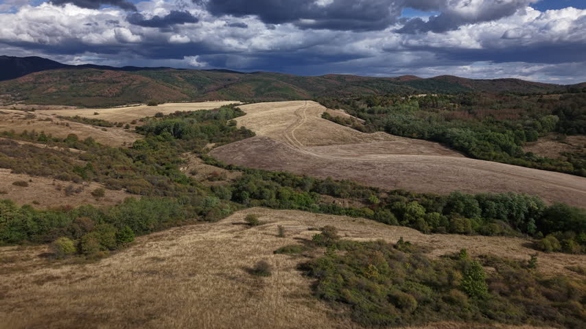 An aerial perspective of rolling golden hills and meadows under a dramatic, cloudy sky, showcasing a vast, serene countryside landscape.