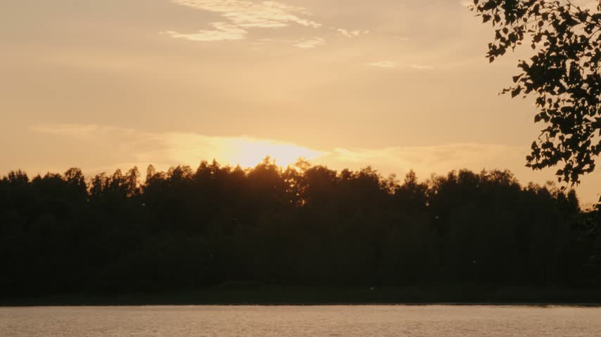 Picturesque sunset glows above a serene lake, casting warm golden light on calm water and forest silhouettes. Peaceful evening landscape in Finland.