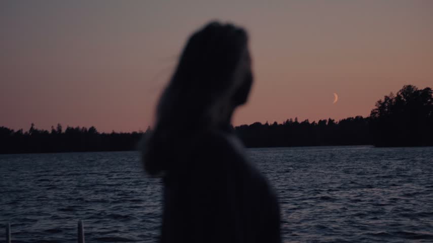 A woman stands by a calm lake at dusk, stretching her arms in silhouette as the crescent moon rises over the water. Warm twilight hues create a peaceful, reflective mood in this cinematic scene.