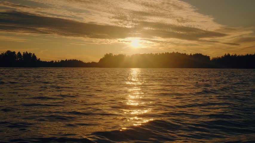 Golden sunset casts warm light over calm Finnish lake, vivid reflections shimmering on water, silhouetted forest horizon, tranquil nature setting.