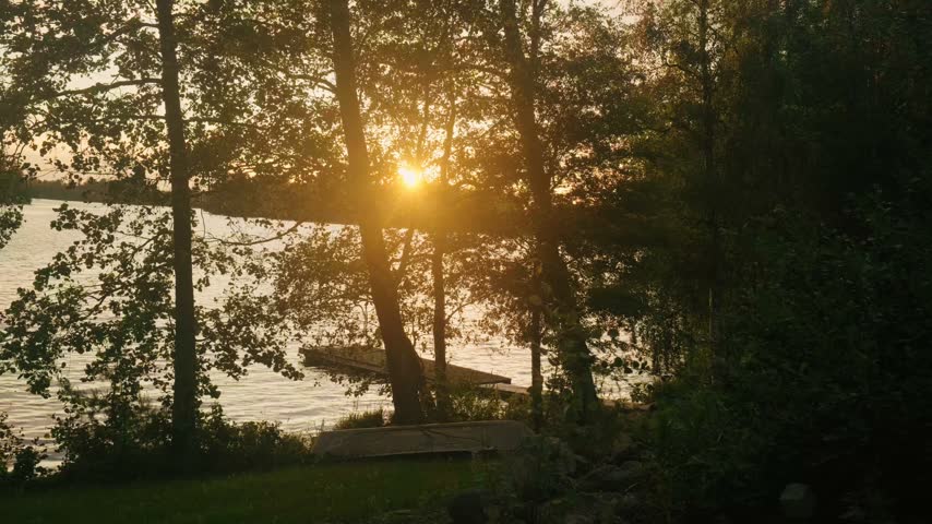 Tranquil sunset over a calm lake in Finland, with warm golden sunlight shining through trees, scenic forested shoreline, wooden dock, and gentle rippling reflections, shot in landscape format.