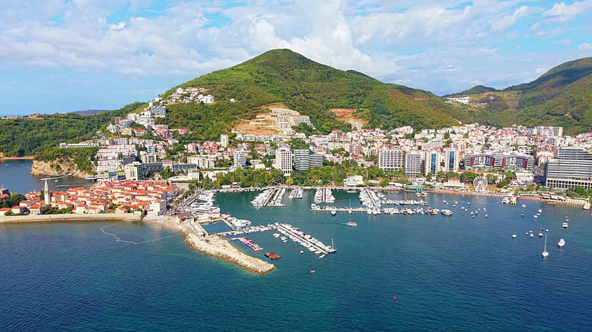Budva, Montenegro rises above Adriatic marina, with dense waterfront buildings framed by green mountain slopes. Aerial panorama tracks harbor, boats and sunlit coastal scenery beneath clouds.