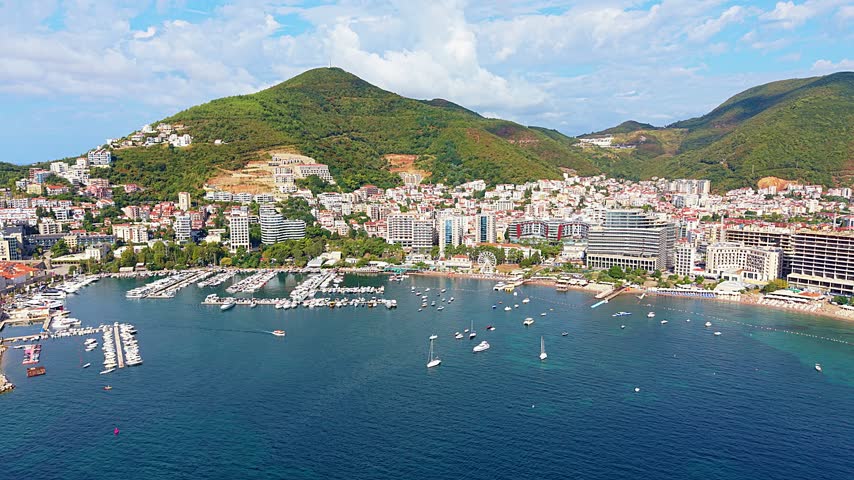 Budva coastal city spreads along Adriatic bay in Montenegro, framed by green mountain slopes. Aerial view tracks across waterfront, marina and anchored boats beneath partly cloudy sky.