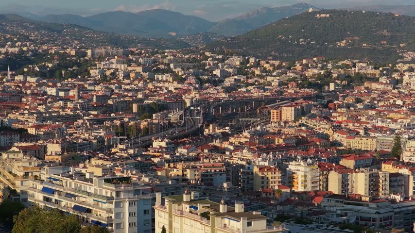 Slow telephoto orbit over apartment blocks and red rooftops toward the rail station and green hills, warm evening light, Nice, French Riviera, Cote d Azur, France