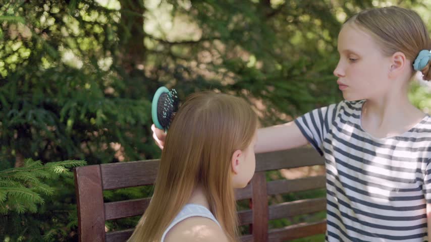 Two sisters share quiet grooming moment sitting together outdoors on park bench. Caring child carefully detangles long blonde hair of younger sibling using plastic hairbrush with gentle care.