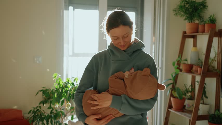 Young smiling mother lovingly holding her cute newborn baby in a cozy brown jumpsuit with bear ears, rocking the infant to sleep in a sun drenched room full of houseplants