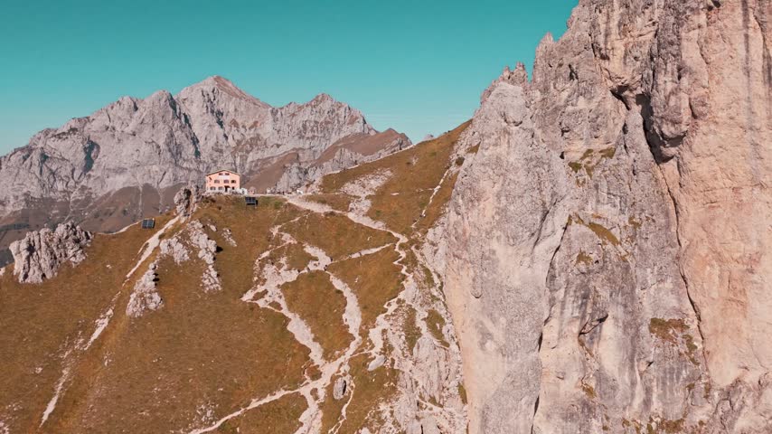 Impressive aerial perspective panning across a rugged mountain landscape, revealing a steep cliff and a mountain hut on a sunny day