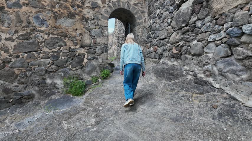Blonde woman wearing a denim jacket and jeans walking up a stone path and through a gate into the courtyard of an ancient medieval castle with a tower, exploring the historical landmark