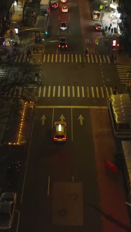 Vertical video: Forwards tracking of typical yellow cab driving through streets of night city. High angle view of taxi passing road intersection. Manhattan, New York City, USA