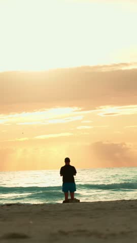 Silhouette of a pensive man watching the sunrise over the ocean, contemplating life and finding inner peace on a serene and tranquil morning beach getaway. Vertical video