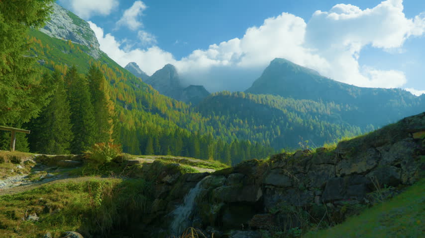 Mountain peaks pine forest autumn landscape with a small water cascade flowing over stones in the foreground. Scenic alpine wilderness scenery with high rocky summits partially covered by white clouds