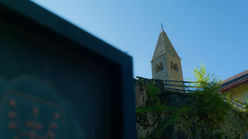 Christian crosses church bell tower standing on a rocky hill against a clear blue sky, handheld panning shot. Low angle view of historic religious calvary monument in alpine environment.