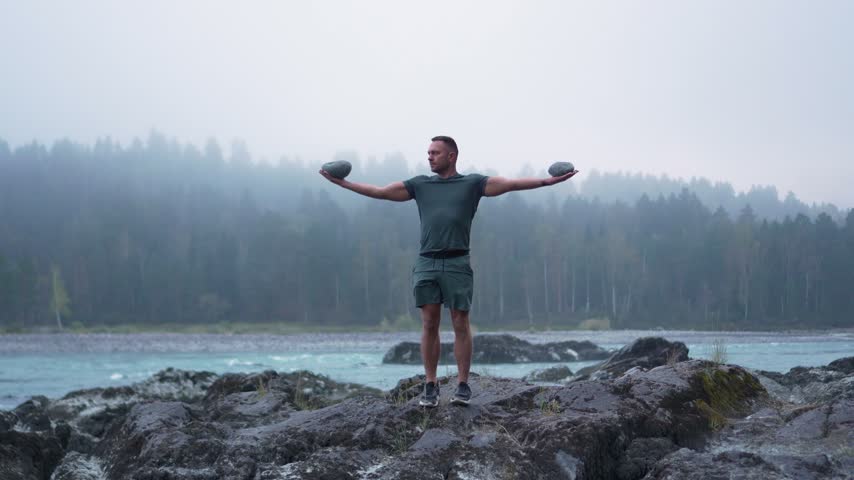 Front view of an athletic man standing on river rocks with arms outstretched holding stones against a foggy forest