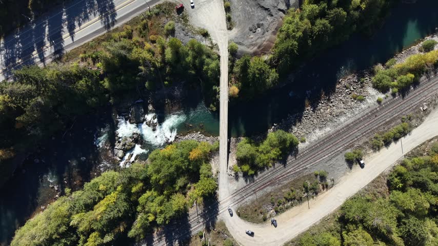 Hovering top down aerial over a bridge crossing a forested river gorge with turquoise water and white rapids surrounded by dense trees, Squamish, British Columbia, Canada.