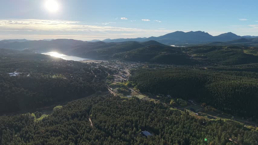Slow side tracking shot over dense pine forest with the mountain town of Nederland and Barker Reservoir in the distance under afternoon sun, Nederland, Boulder County, Colorado