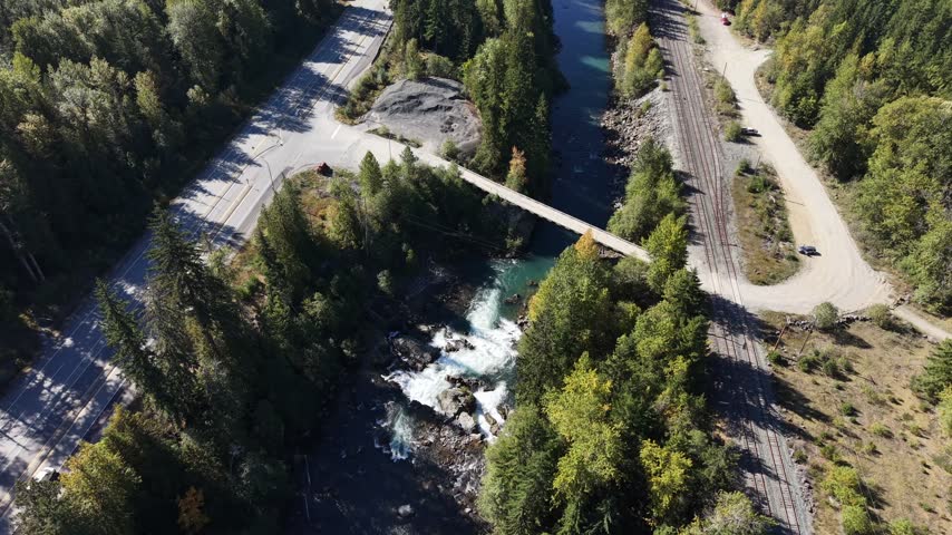 Panoramic top down aerial hold over a bridge crossing a forested river gorge with white water rapids and a road visible through the trees, Squamish, British Columbia, Canada.