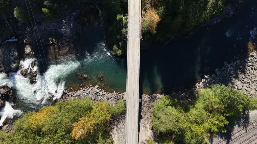 High angle static aerial over a bridge spanning a deep canyon with turquoise water and white rapids flanked by dense forest with autumn foliage, Squamish, British Columbia, Canada.