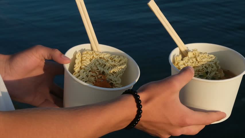 Young adult hands hold two cups of instant noodles with chopsticks before adding hot water, representing a quick outdoor meal after hiking with camping food, convenient meals, and travel nutrition