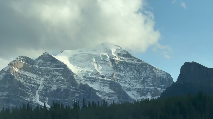 A handheld shot captures the majestic Banff National Park, where towering pine trees partially obscure the rugged mountain peaks under a partly cloudy sky, highlighting the serene beauty of the Canadi