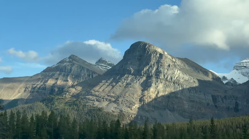 A serene view of the Rocky Mountains under a vast, clear blue sky, with patches of scattered clouds adding depth to the scene. The dense forest at the base of the mountains provides a lush foreground,
