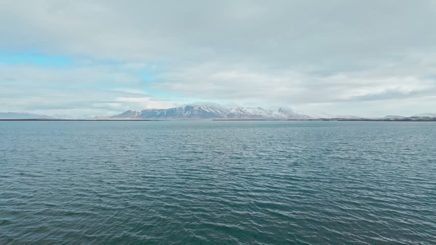 A landscape view of Mt Esja in Iceland behind a foreground of a vast and open bay of blue water under a bright cloudy sky.