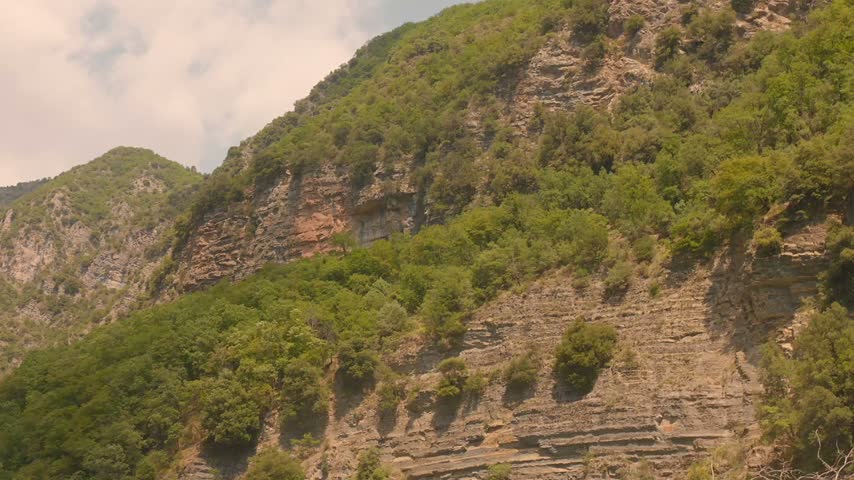 Rocky green mountainside under partly cloudy sky, showing natural layered cliffs