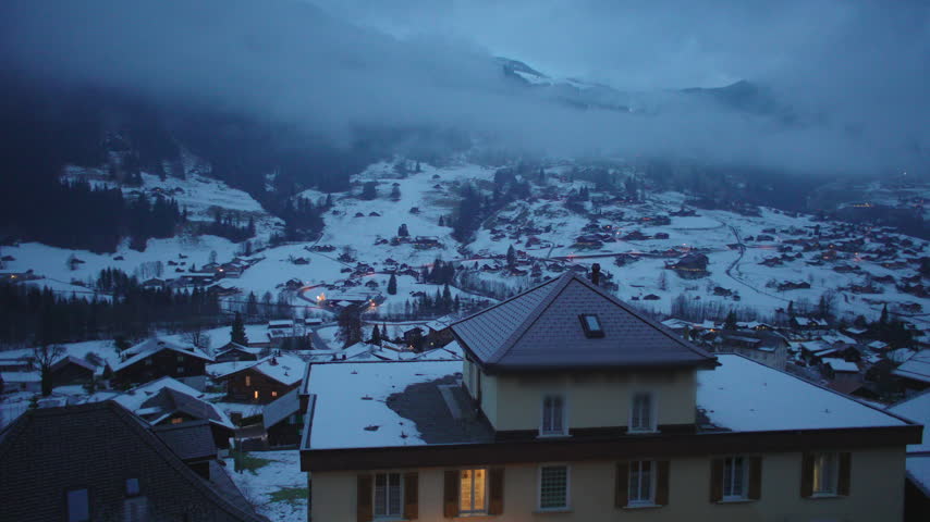 Elevated view of small town surrounded by snowy hills and misty peaks, quiet residential houses and winding roads under dramatic winter sky